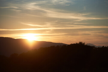 Sun sets behind rolling hills, casting warm, golden glow across sky. Wispy clouds, tinged with hues of orange and yellow, stretch across deepening blue sky. Serene and expansive landscape.