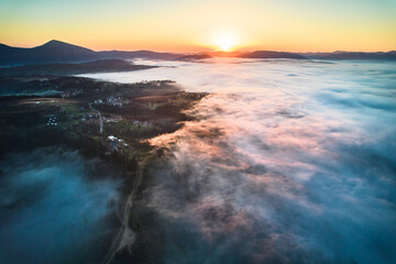 Stunning aerial view of sunrise over mountainous landscape, with thick fog blanketing valleys and hills. Sun's golden rays pierce through mist, casting serene glow over the entire scene.