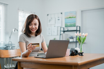 A young woman using a credit card to shop online with a laptop in a modern home office, surrounded by decor and natural light.
