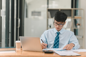 Fototapeta premium A man wearing a blue tie is sitting at a desk with a laptop and a stack of papers. He is writing on the papers with a pen. The scene suggests a professional setting