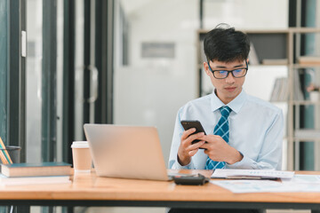 A man in a blue shirt and blue tie is sitting at a desk with a laptop and a cell phone