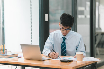 Fototapeta premium A man in a blue shirt and tie is sitting at a desk with a laptop and a calculator. He is writing on a piece of paper and he is focused on his work. Concept of professionalism and productivity