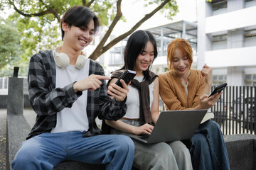 Fototapeta premium Three young asian people happily using technology outdoors, connecting with friends and the community through smartphones and laptops, embodying togetherness and youth in the digital age