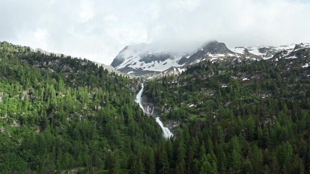 Scenic view of Rutor Waterfalls cascading down a lush green forest with snowy mountains in the background