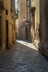 Fototapeta premium Dettaglio di un vicolo di Lecce con un raggio di sole che colpisce un lampione e proietta la sua ombra sul selciato