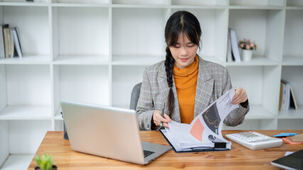 A positive Asian businesswoman is reviewing the financial report while working at her desk.