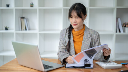 A positive Asian businesswoman is looking at the laptop screen while reviewing financial reports.