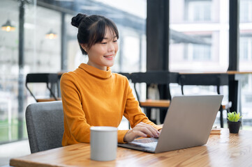 A charming Asian woman is working remotely from a coffee shop, working on her laptop computer.
