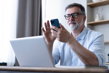 Handsome smiling senior man wearing glasses using mobile phone while sitting at his workplace with laptop at home