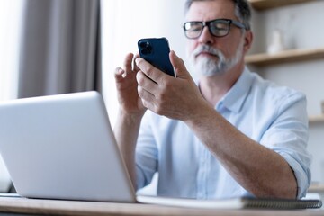 Handsome smiling senior man wearing glasses using mobile phone while sitting at his workplace with laptop at home