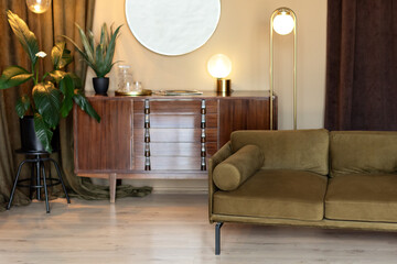Mid century room in home with modern interior design, green velvet sofa, wooden cabinet, mirror, houseplants in pots and loft pendant lamps. Living room with furniture. Retro Edison light bulbs. 