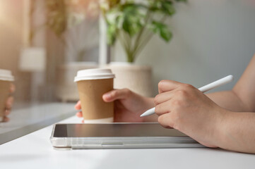 A woman holding a stylus pen, writing on a digital tablet, working remotely from a coffee shop.