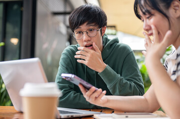 An excited, stunned young Asian man is surprised with the news on his friend's smartphone.