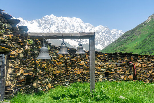 Three bells hang on a wooden log near a stone wall in the courtyard of the Lamaria Monastery in the village of Ushguli. The snow capped peaks of Mount Shkhara are in the background.