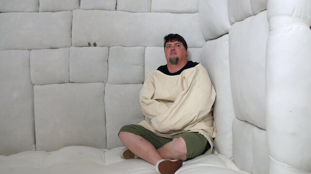 A crazy man in a straitjacket sitting hunched in the corner of a padded cell room, his expression somber. Despair depression in isolation in an insane asylum