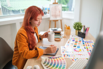 Female graphic designer using a digital tablet at creative workspace surrounded by various colour swatches and design charts.