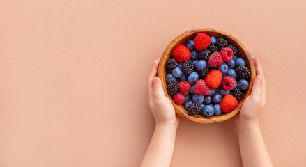 Child hands holding bowl of fresh summer berries on beige background top view. Organic food, snack...