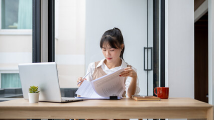 Obraz premium A focused Asian businesswoman is reviewing business report at her desk, working in her office.