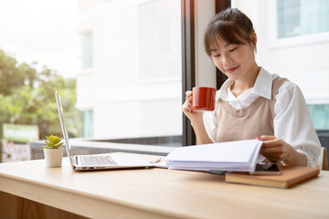 A beautiful Asian businesswoman having a morning coffee while reviewing business report at her desk.