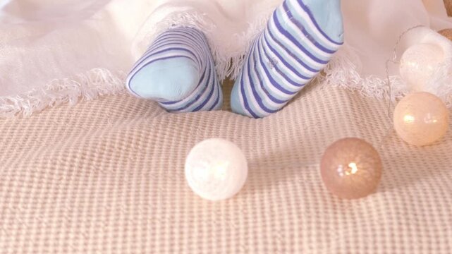 Woman covering herself with blanket, close-up feet in blue socks, dozing on bed in peaceful setting, garland balls for coziness, domestic bliss, relaxation retreat, Cozy Comfort