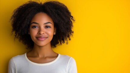 A woman with curly hair is smiling at the camera. She is wearing a white shirt and standing in front of a yellow wall
