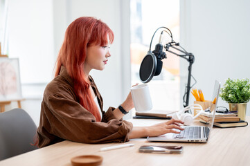 A beautiful Asian female podcaster is working in her home studio and enjoying a coffee.