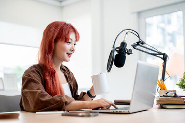 A beautiful Asian female podcaster is working in her home studio and enjoying a coffee.
