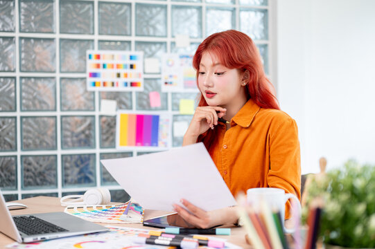 A focused, creative Asian female graphic designer with red hair is working at her desk in the office