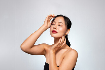 A beautiful Asian woman poses against a grey background, touching her face with her eyes closed.