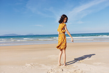 Beautiful woman in a flowing yellow dress standing gracefully on the sandy beach with the tranquil ocean as the backdrop