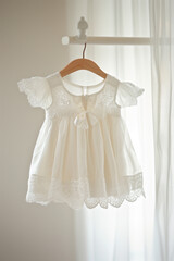 beautifully crafted baby dress featuring a delicate lace overlay and a soft, flowing skirt. The dress is hung on a wooden hanger against a neutral background,