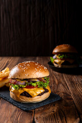 Gourmet cheeseburger with bacon, lettuce, tomato, onion, and melted cheese on a sesame bun, served with fries on a rustic wooden table, dark background. Another burger in the background adds depth