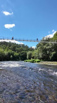 Xirimbao in Ulla river in Galicia. A picturesque suspension bridge spans a flowing river, surrounded by lush greenery and dense forest, under a clear blue sky, capturing the serene beauty of nature.