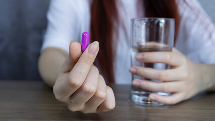 Medicine, Asian woman's hand holding violet pill capsule carefully with fingers and holding glass of water, symbolizing healthcare, Concept of pharmacy, medicine, supplement and medical treatment.