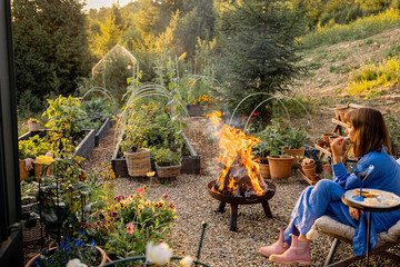 A woman enjoys a peaceful evening by the fire in a lush garden, surrounded by flowers and greenery. The setting sun casts a warm glow, enhancing the tranquil atmosphere of this outdoor retreat