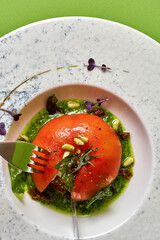 tomato, garnished with pine nuts and herbs, served on a white plate against a green background.