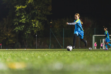 cute little girl with blue uniform kicking a ball at the evening match, full shot. High quality...
