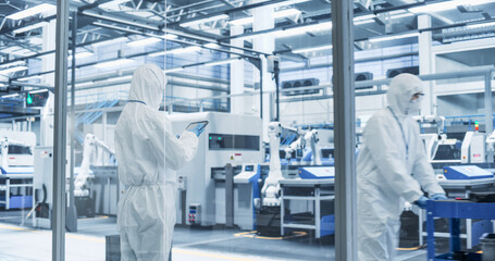 Industrial Scientist Wearing Disposable Protective Suit to Be Protected From Chemical and Biological Dangers. Female Using Tablet, Monitoring Production Process at a Biotechnology Plant