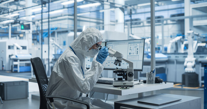 Research Factory Cleanroom: Engineer Wearing Coveralls, Gloves, and Goggles while Using Microscope to Inspect a Microchip. Scientist Developing an Advanced Electronics Component for Precision Industry