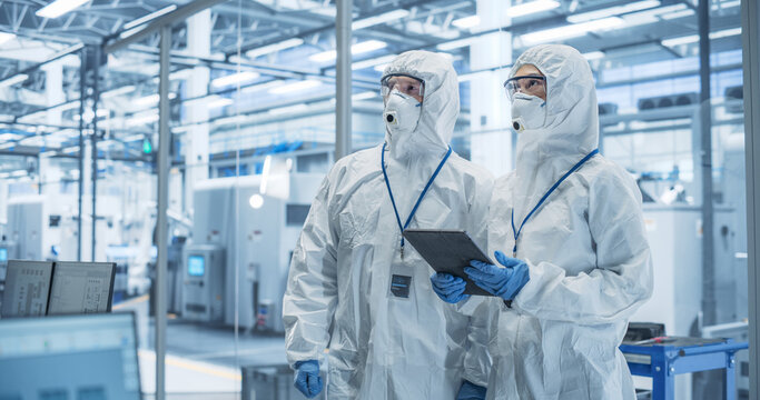 Industrial Scientists Wearing Disposable Protective Suits, Masks and Goggles at Production Plant. Specialist Using Tablet Computer, Analyzing Potential Work Hazards and Contamination at a Workplace