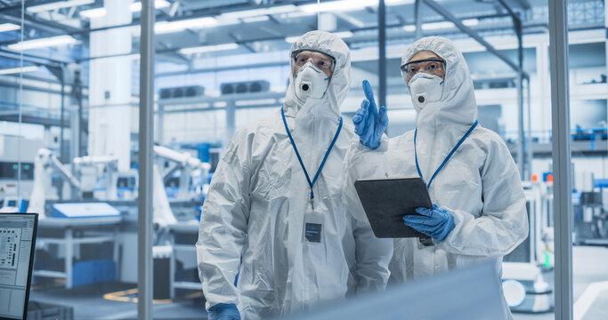 Industrial Scientists Wearing Disposable Protective Suits, Masks and Goggles at a Production Plant. Specialist Using Tablet Computer, Analyzing Potential Work Hazards and Contamination at a Workplace