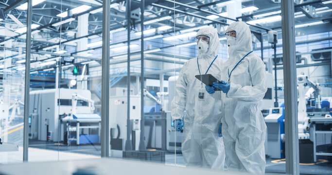 Development Factory Cleanroom: Engineers Wearing Coveralls and Goggles while Talking and Using a Tablet Computer. Scientists Working in Advanced Electronics Manufacture with Precision Machinery