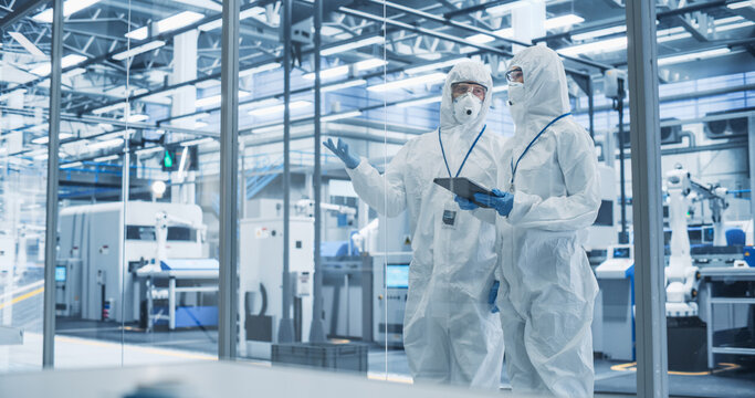 Development Factory Cleanroom: Engineers Wearing Coveralls and Goggles while Talking and Using Tablet Computer. Scientists Working in Advanced Electronics Manufacture with Precision Machinery