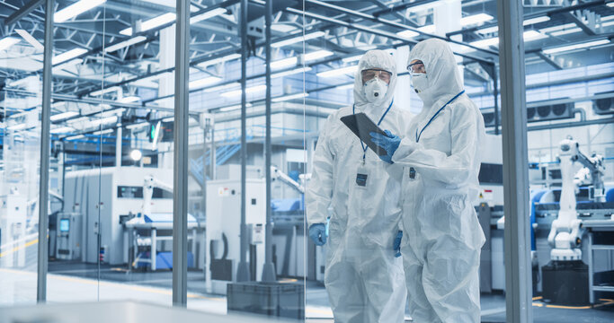 Development Factory Cleanroom: Engineers Wearing Coveralls and Goggles while Talking and Using Tablet. Scientists Working in Advanced Electronics Manufacture with Precision Machinery