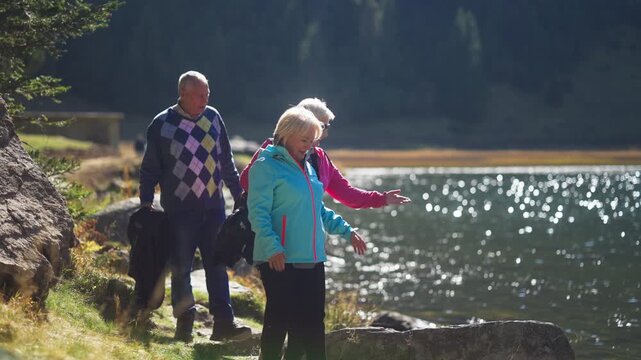 Two Elderly Caucasian Women and a Man Exploring Nature Together and Walking by a Mountain Lake on a Sunny Autumn Day. Concept of weekend activity, healthy lifestyle, vitality, retirement, well-being