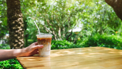 A hand holding a glass of iced latte coffee on wooden table in the garden