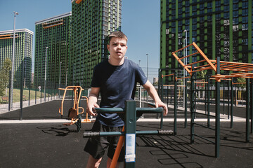 A boy on a sports ground trains on outdoor exercise machines. Legs and thighs training.