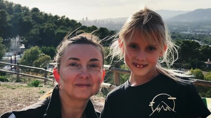 Portrait of smiling mother and cute daughter in the natural park. Middle-aged caucasian woman hugging with 7 years old blonde girl looking at camera. City skyline and mountains on background.
