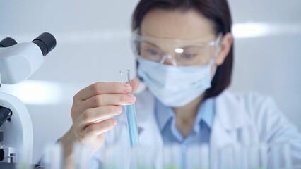 Scientist female researcher with protective glasses and mask examining lab tube with a blue fluid. Microbiology and science concept