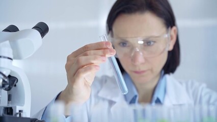 Scientist female researcher with protective glasses examining lab tube with a blue fluid. Microbiology and science concept